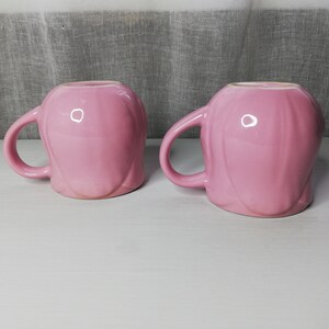 May include: Two pink ceramic mugs with a smooth, rounded design. The mugs have a handle on one side and are perfect for enjoying a warm beverage.