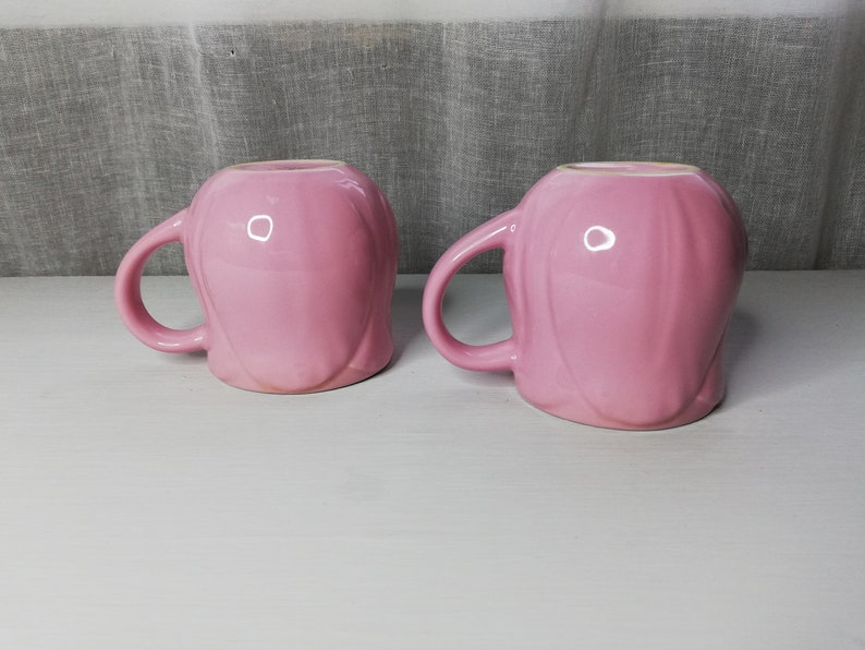 May include: Two pink ceramic mugs with a smooth, rounded design. The mugs have a handle on one side and are perfect for enjoying a warm beverage.