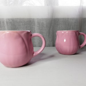 May include: Two pink ceramic mugs shaped like tulips with a white interior. The mugs have a handle on the right side.