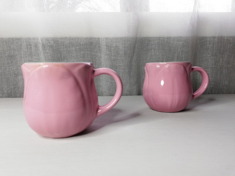 May include: Two pink ceramic mugs shaped like tulips with a white interior. The mugs have a handle on the right side.