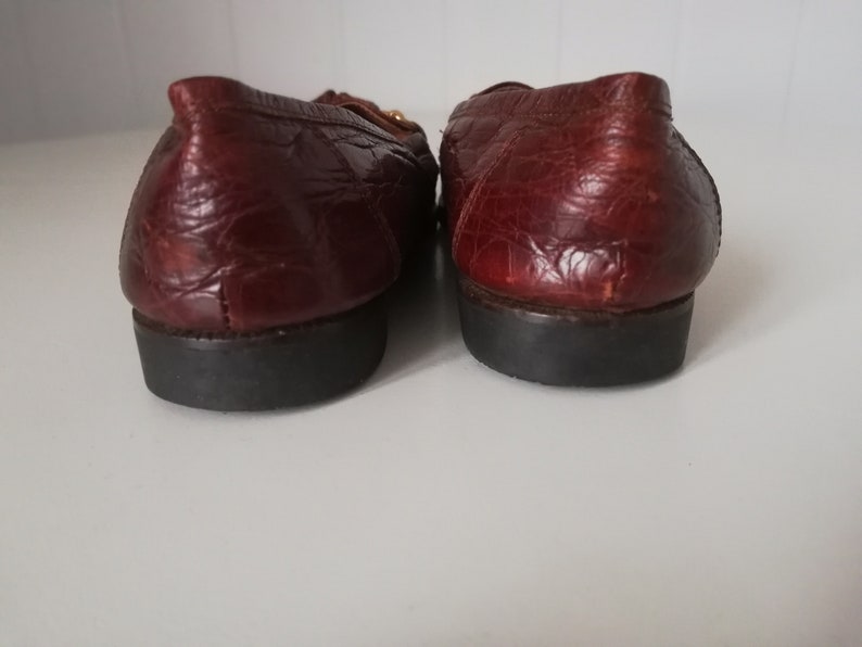 Puede incluir: Un par de mocasines de cuero marr&oacute;n con un patr&oacute;n de cocodrilo texturizado. Los mocasines tienen un detalle de hebilla dorada y una suela de goma negra.