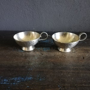 May include: Two silver hammered metal cups with handles. The cups are sitting on a dark wooden surface.