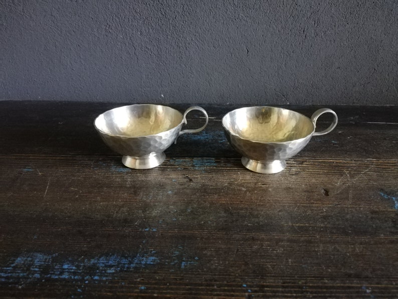 May include: Two silver hammered metal cups with handles. The cups are sitting on a dark wooden surface.