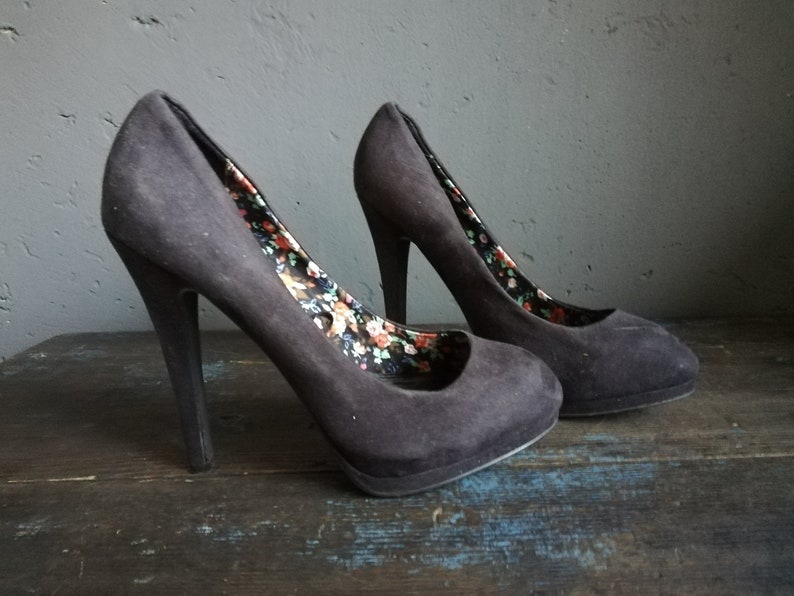 Op de afbeelding: Een paar zwarte su&egrave;de pumps met een bloemenprint voering. De hakken zijn ongeveer 10 cm hoog.