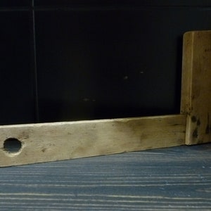 May include: A wooden carpenter's square with a hole in the longer arm. The square is made of light brown wood and has a worn, rustic appearance.