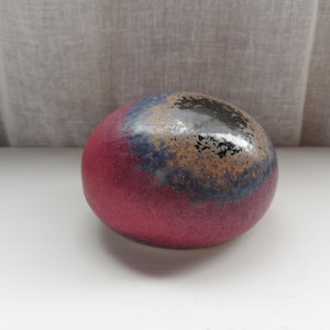 Op de afbeelding: Een glad, rond papieren gewicht van keramiek met een roze, blauwe en zwarte glazuur. De glazuur heeft een gespikkeld effect, waardoor een unieke en artistieke uitstraling ontstaat.