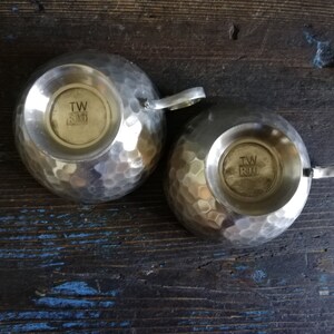 May include: Two silver hammered metal cups with handles. The bottom of each cup has a circular stamp with the text "TW & RIO".