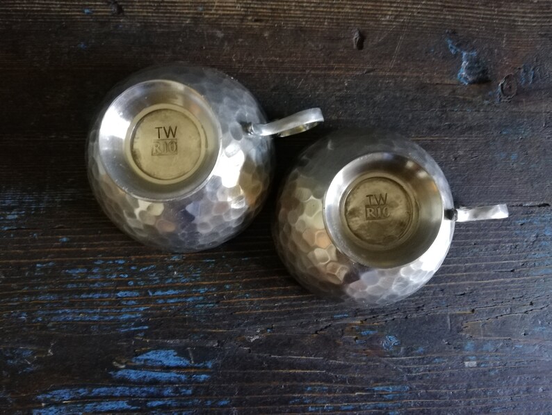 May include: Two silver hammered metal cups with handles. The bottom of each cup has a circular stamp with the text "TW & RIO".