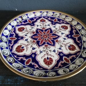 May include: A decorative ceramic plate with a white background and a blue and red floral design. The plate has a gold rim and a white and black swirl pattern around the edge.