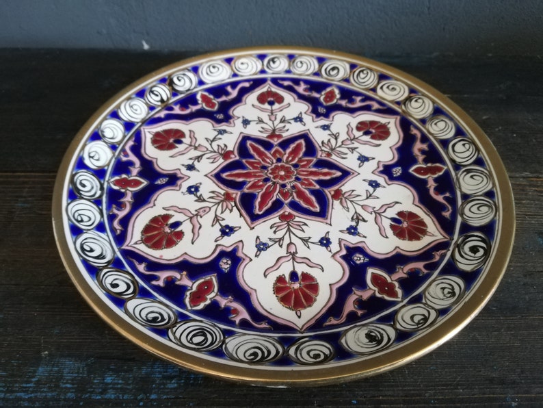 May include: A decorative ceramic plate with a white background and a blue and red floral design. The plate has a gold rim and a white and black swirl pattern around the edge.