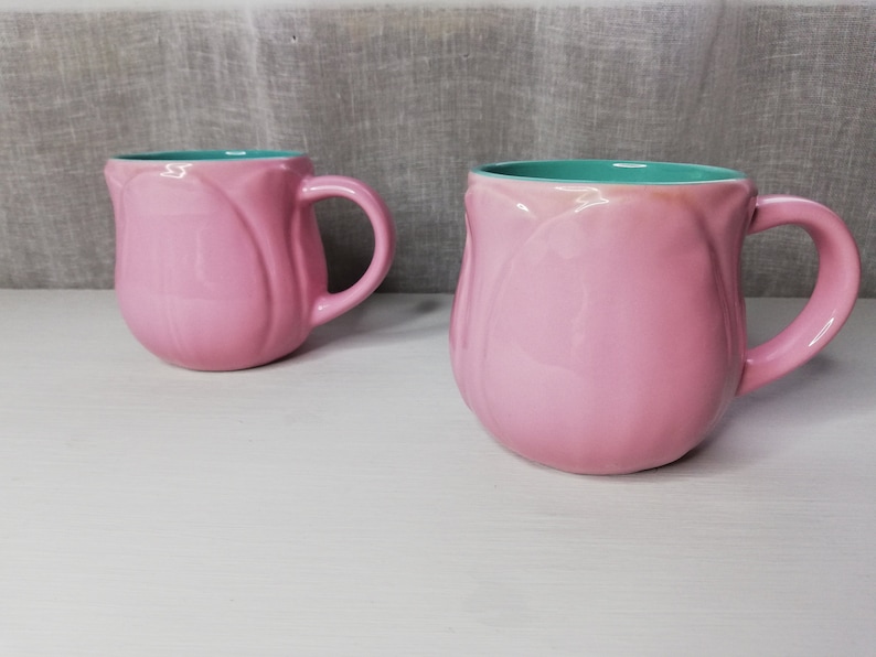 May include: Two pink ceramic mugs with a light blue interior. The mugs are shaped like tulips and have a rounded handle.