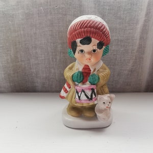 Puede incluir: Una figurita de cerámica de un niño que lleva un gorro rojo y blanco, un abrigo verde y marrón, y sostiene un tambor rosa y blanco. El niño está de pie junto a un pequeño cerdo blanco.