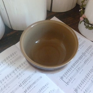 May include: A ceramic bowl with a brown interior and a tan exterior with brown stripes. The bowl is sitting on a sheet of music. The bowl is a kitchenware item.