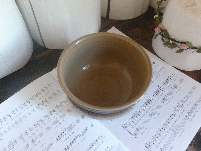 May include: A ceramic bowl with a brown interior and a tan exterior with brown stripes. The bowl is sitting on a sheet of music. The bowl is a kitchenware item.