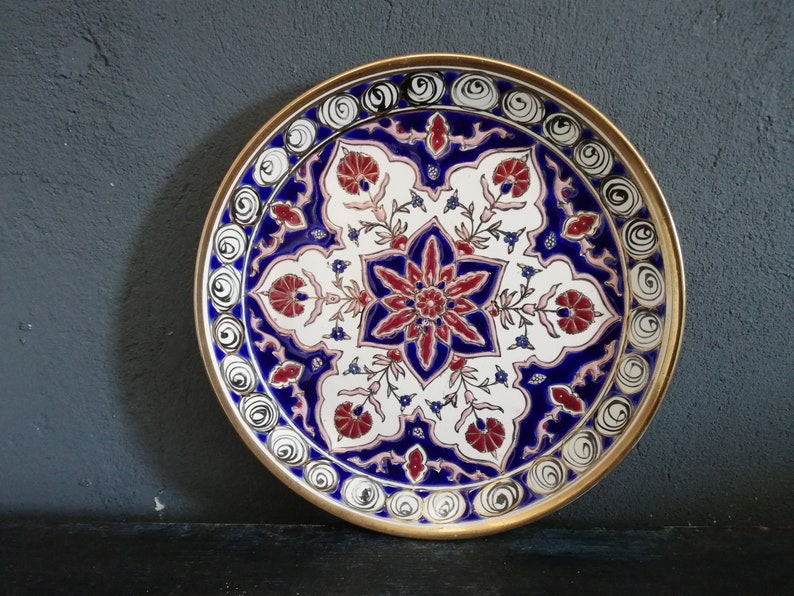 May include: A round decorative plate with a gold rim and a white background. The plate features a blue and red floral design with intricate details.