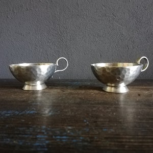 May include: Two silver hammered metal cups with handles. The cups have a rounded bottom and a raised base.