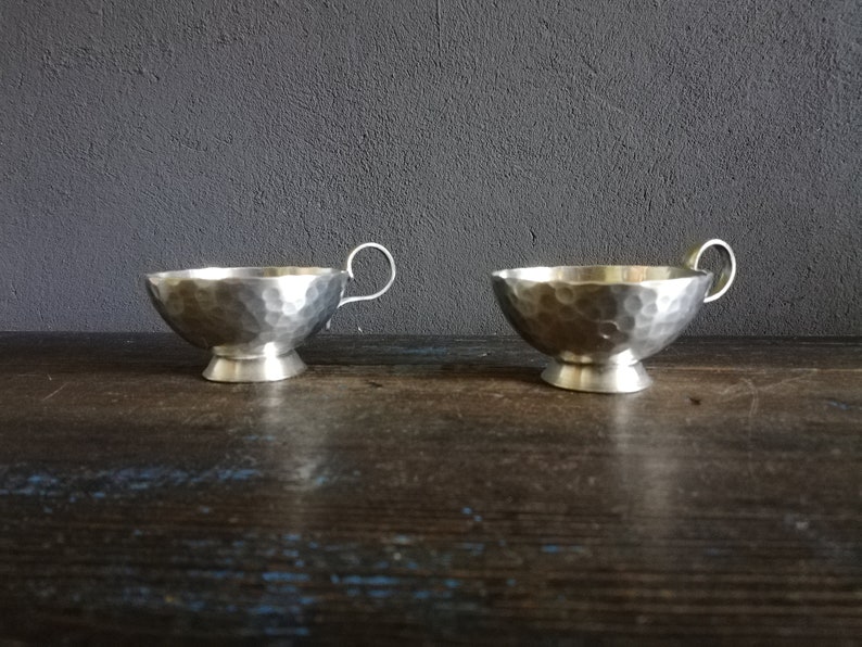 May include: Two silver hammered metal cups with handles. The cups have a rounded bottom and a raised base.