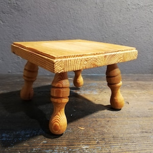 Puede incluir: Un pequeño taburete de madera con cuatro patas torneadas. El taburete es de madera marrón claro y tiene una tapa cuadrada.