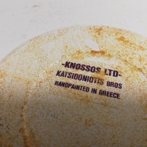Puede incluir: Primer plano de un plato de cer&aacute;mica blanco con una mancha marr&oacute;n. El plato tiene un sello morado que dice "KNOSSOS LTD - KATSIDONICTS BROS HANDPAINTED IN GREECE".