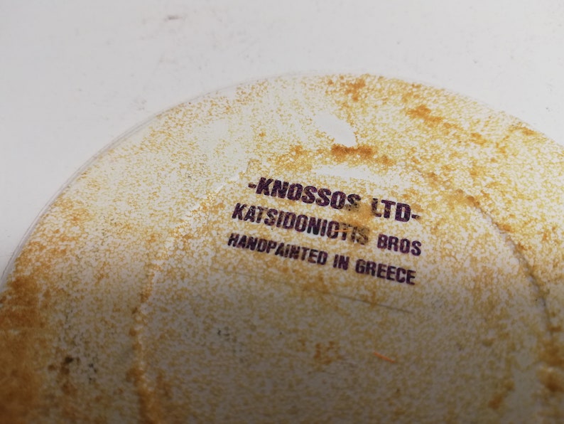 Puede incluir: Primer plano de un plato de cer&aacute;mica blanco con una mancha marr&oacute;n. El plato tiene un sello morado que dice "KNOSSOS LTD - KATSIDONICTS BROS HANDPAINTED IN GREECE".