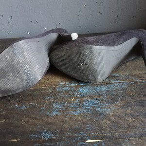 Op de afbeelding: Een paar zwarte su&egrave;de pumps met hoge hakken en een spitse neus. De schoenen liggen op een houten oppervlak.