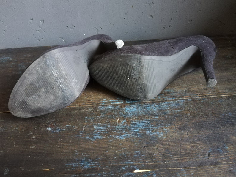 Op de afbeelding: Een paar zwarte su&egrave;de pumps met hoge hakken en een spitse neus. De schoenen liggen op een houten oppervlak.