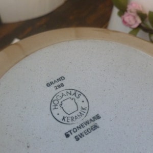 May include: A beige stoneware plate with a speckled interior and a light brown rim. The plate features the text "GRAND 298" and a circular logo with the words "HOGANAS KERAMIK STONEWARE SWEDEN".
