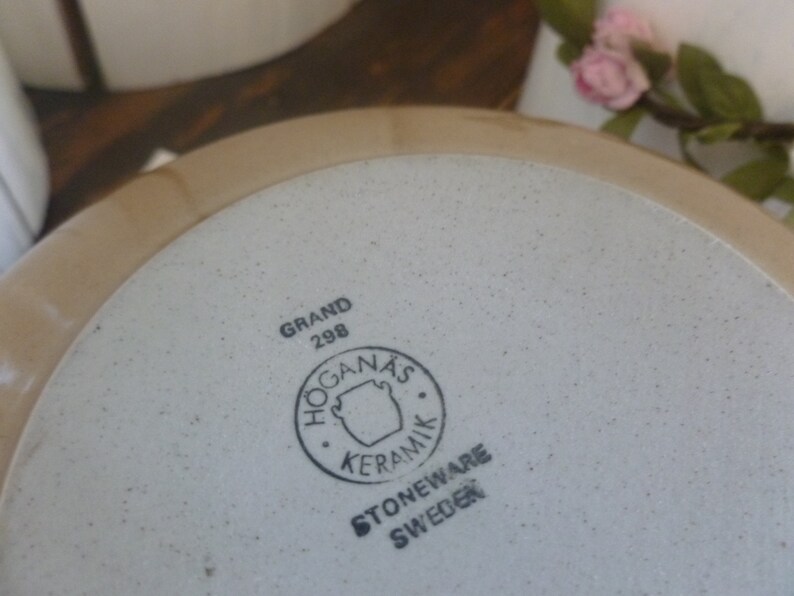 May include: A beige stoneware plate with a speckled interior and a light brown rim. The plate features the text "GRAND 298" and a circular logo with the words "HOGANAS KERAMIK STONEWARE SWEDEN".
