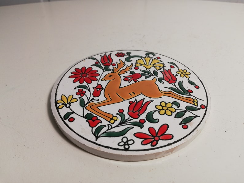 Puede incluir: Un posavasos de cer&aacute;mica blanca con un dise&ntilde;o floral colorido que presenta un ciervo en el centro. El ciervo es naranja y marr&oacute;n con detalles blancos. Las flores son rojas, amarillas y verdes.