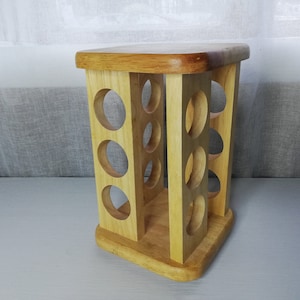 Puede incluir: Un estante para especias de madera con 12 aberturas redondas para guardar especias. El estante está hecho de madera marrón claro y tiene una base cuadrada.