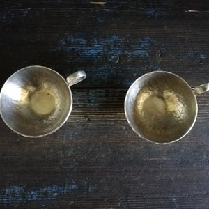 May include: Two silver cups with hammered metal surfaces and handles. The cups are empty and sitting on a dark wooden surface.