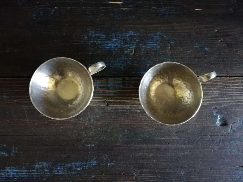 May include: Two silver cups with hammered metal surfaces and handles. The cups are empty and sitting on a dark wooden surface.