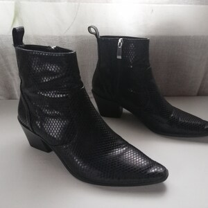 Może przedstawiać: Para czarnych sk&oacute;rzanych kowbojskich but&oacute;w ze spiczastym noskiem i wzorem sk&oacute;ry węża. Buty mają obcas blokowy i zamek błyskawiczny po wewnętrznej stronie kostki.