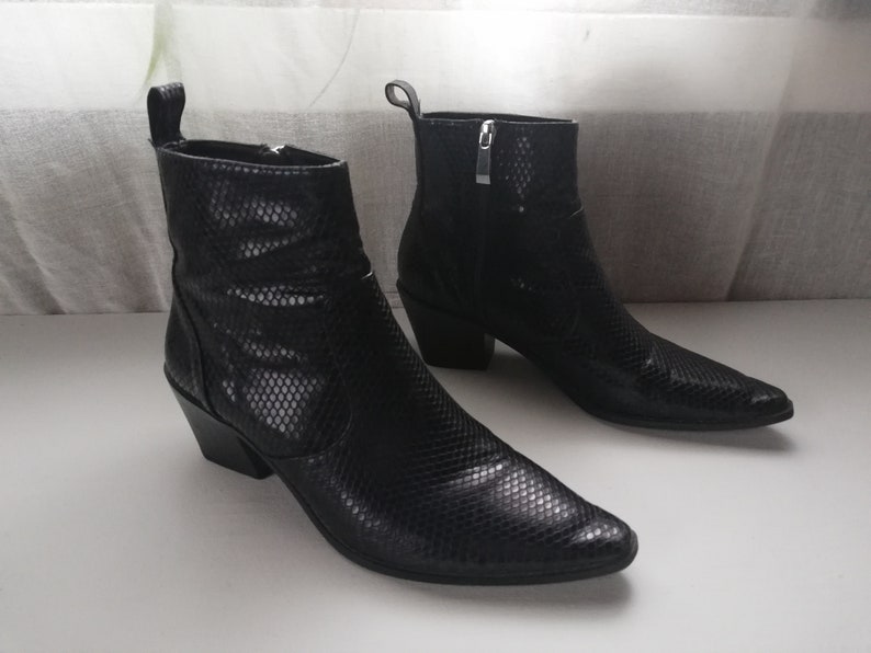 Może przedstawiać: Para czarnych sk&oacute;rzanych kowbojskich but&oacute;w ze spiczastym noskiem i wzorem sk&oacute;ry węża. Buty mają obcas blokowy i zamek błyskawiczny po wewnętrznej stronie kostki.