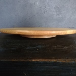 Puede incluir: Un soporte para tartas redondo de madera con un acabado de madera natural. El soporte está sobre una superficie azul oscuro.