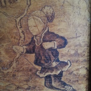 May include: A woodburned image of a person wearing a hat and coat, holding a rope, with a tree in the background.