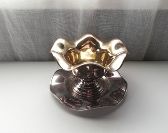 May include: A silver and gold serving dish with a scalloped edge and a pedestal base. The dish is designed for serving sauces or other small dishes.