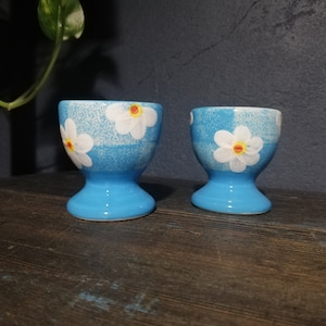 May include: Two ceramic egg cups. The cups are light blue with white daisy floral designs and a yellow center. The egg cups have a pedestal base and are displayed on a dark wooden surface.