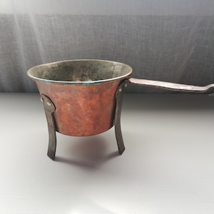 Puede incluir: Una pequeña olla de cobre con asa y tres patas. La olla está hecha de cobre y tiene un acabado de pátina. El asa está hecha de hierro y tiene un gancho en el extremo.