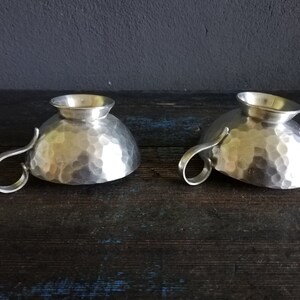 May include: Two silver hammered metal cups with handles. The cups are sitting on a dark wooden surface.