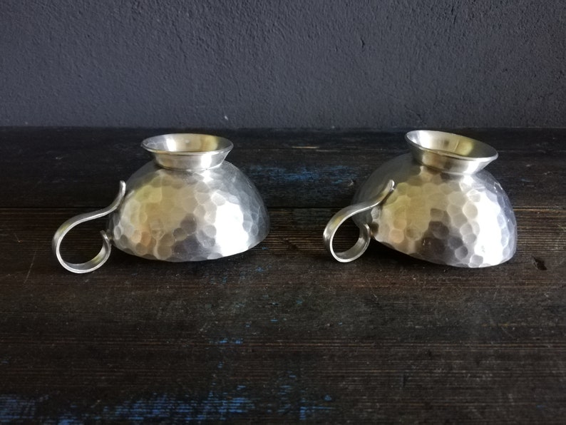 May include: Two silver hammered metal cups with handles. The cups are sitting on a dark wooden surface.
