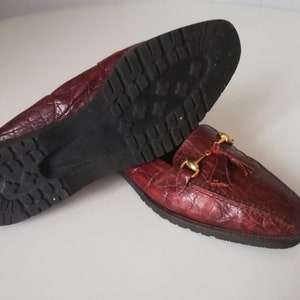 Puede incluir: Un par de mocasines de cuero burdeos con un borla y herrajes dorados. Los mocasines tienen una parte superior de cuero texturizado y una suela de goma.
