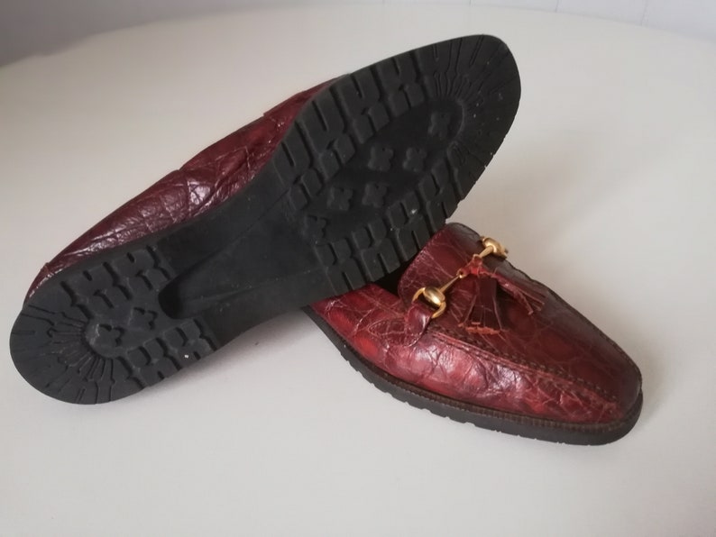 Puede incluir: Un par de mocasines de cuero burdeos con un borla y herrajes dorados. Los mocasines tienen una parte superior de cuero texturizado y una suela de goma.