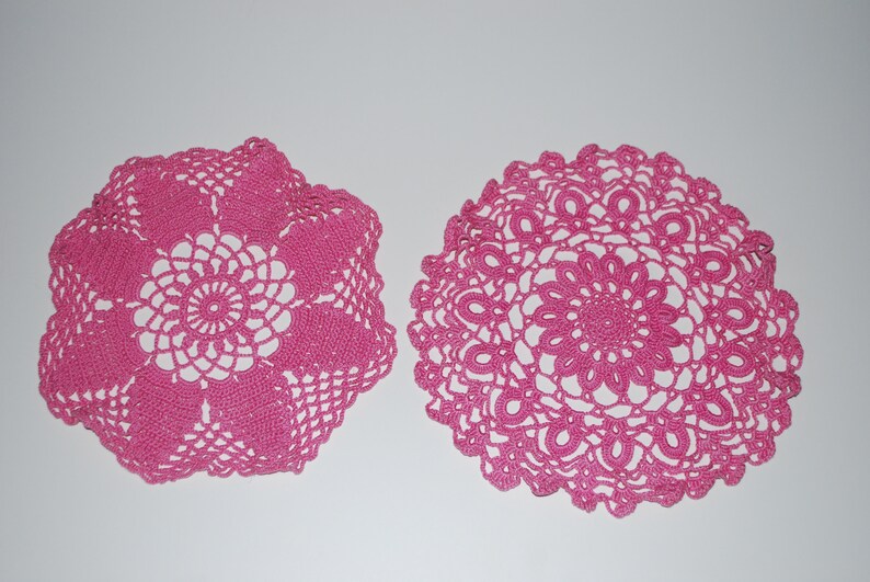 May include: Two pink crocheted doilies, one with a circular design and the other with a square design.
