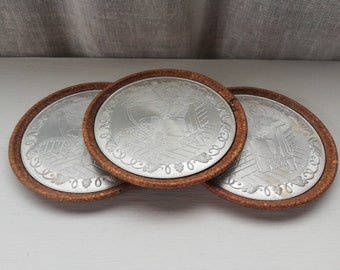 May include: Three round coasters with a brown cork-like outer rim and a silver metal center. Each coaster features a different design of a person in a medieval-style outfit.