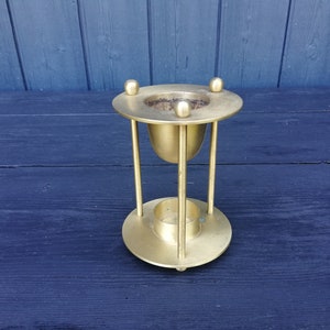 May include: A vintage brass incense burner with a round base and top, connected by vertical supports. The top features three small decorative spheres. The burner has a cup-shaped interior and a small circular base for a candle.