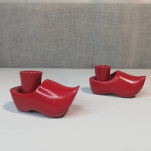Op de afbeelding: Twee rode houten kaarsenhouders in de vorm van klompen. De kaarsenhouders zijn gemaakt van hout en hebben een gladde, glanzende afwerking.