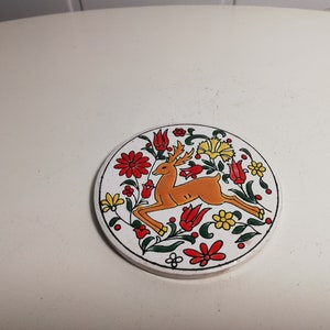 Puede incluir: Un posavasos de cer&aacute;mica blanca con un dise&ntilde;o floral colorido que presenta un ciervo en el centro. El ciervo es naranja y marr&oacute;n con contornos negros. Las flores son rojas, amarillas y verdes.