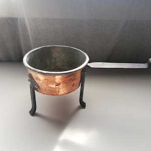 Op de afbeelding: Een kleine koperen pot met een zwart metalen handvat en drie poten. De pot staat op een wit oppervlak.
