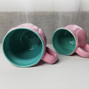 May include: Two ceramic mugs with pink exteriors and teal interiors. The mugs have a rounded shape and a handle on the side.